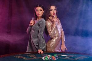 Two girls are playing poker at casino, holding glasses of champagne, posing at a table with chips, cards on it. Black, smoke background. Close-up. photo