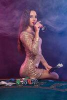 Girl playing poker at casino, holding money, drinking champagne. Sitting on table with chips, cash on it. Black, smoke background. Close-up. photo