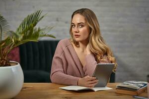 Female blogger in pink cardigan is holding a tablet. Sitting at wooden table with notebooks and palm tree in pot on it. Student, blogger. Close-up photo