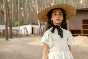 Cute dark haired preteen girl wearing light blouse and large brim straw hat posing on country estate in pine forest on summer day, looking confidently at camera photo