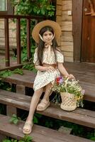 Smiling dark-haired preteen girl sitting on doorstep of rural cottage with basket of wildflowers photo