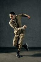 Modern guy with tattooed body and face, earrings, beard. Dressed in khaki overalls and black sneakers. Dancing on gray background. Dancehall, hip-hop photo