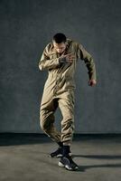 Strong performer with tattooed body, earrings, beard. Dressed in khaki overalls and black sneakers. Dancing on gray background. Dancehall, hip-hop photo