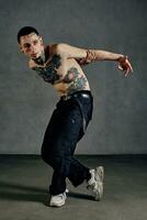 Athletic tattooed guy with naked torso, beard. Dressed in black pants and white sneakers. Dancing against gray background. Dancehall, hip-hop photo