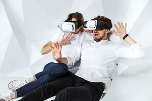 A young couple wearing VR headsets sitting on white chair in a room with white walls and floors photo