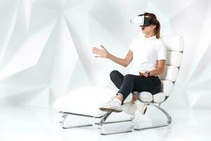 Technology, virtual reality, entertainment and people concept - happy young woman with virtual reality headset sits on a white armchair photo