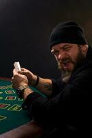 Serious bearded man sitting at poker table and holding cards isolated on black. Copy space photo