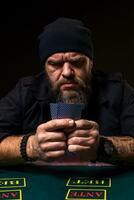 Serious bearded man sitting at poker table and holding cards isolated on black photo