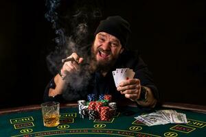 hombre es jugando póker con un cigarro y un whisky, un hombre espectáculo dos tarjetas en el mano, victorioso todas el papas fritas en el mesa con grueso cigarrillo fumar. foto