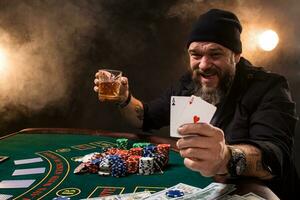 hombre es jugando póker con un cigarro y un whisky, un hombre espectáculo dos tarjetas en el mano, victorioso todas el papas fritas en el mesa con grueso cigarrillo fumar. foto