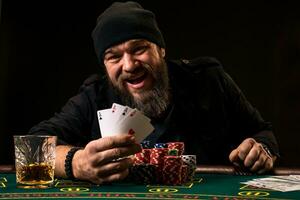 barbado hombre con cigarro y vaso sentado a póker mesa y gritando aislado en negro foto