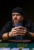 Serious bearded man sitting at poker table and holding cards isolated on black photo