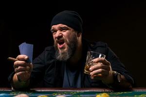 Bearded man with cigar and glass sitting at poker table and screaming isolated on black photo