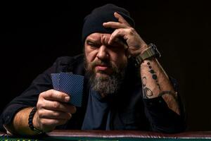 Serious bearded man sitting at poker table and holding cards isolated on black photo