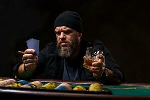 grave barbado hombre con cigarro y vaso sentado a póker mesa y gritando aislado en negro foto