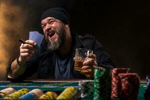 Bearded man with cigar and glass sitting at poker table and screaming isolated on black photo