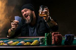 Bearded man with cigar and glass sitting at poker table and screaming isolated on black photo
