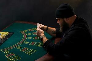 Serious bearded man sitting at poker table and holding cards isolated on black. Copy space photo