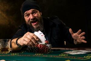 Bearded man with cigar and glass sitting at poker table and screaming isolated on black photo