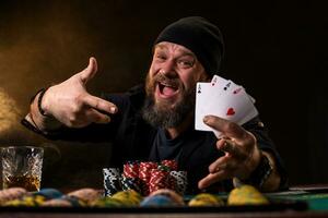 Bearded man with cigar and glass sitting at poker table and screaming isolated on black photo