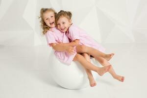 Two little girlfriends in the same pink dresses sitting on a chair in a studio with white walls photo