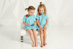 Two little girlfriends in the same turquoise dresses sitting on a chair in a studio with white walls photo