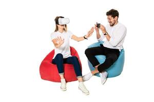 Smiling friends experiencing virtual reality glasses seated on beanbags isolated on white background. photo