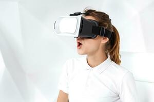 Technology, virtual reality, entertainment and people concept - happy young woman with virtual reality headset sits on a white armchair photo