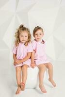 Two little girlfriends in the same pink dresses sitting on a chair in a studio with white walls photo