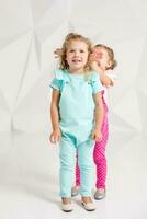 Two little girlfriends in the identical overalls of different colors in a studio with white walls photo