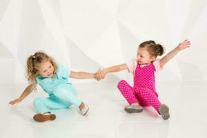 Two little girlfriends in the identical overalls of different colors sitting on the floor in a studio with white walls photo