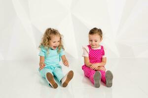 Two little girlfriends in the identical overalls of different colors sitting on the floor in a studio with white walls photo