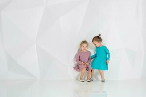 Two little girlfriends in the identical dresses of different colors sitting in a studio with white walls photo