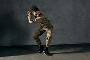 Graceful guy with tattooed body and face, earrings, beard. Dressed in khaki overalls, black sneakers. Dancing on gray background. Dancehall, hip-hop photo