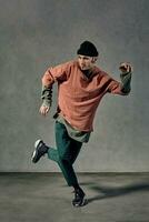 Strong man with tattooed body, earrings, beard. Dressed in hat, casual clothes and black sneakers. Dancing on gray background. Dancehall, hip-hop photo