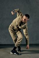 Flexible guy with tattooed body and face, earrings, beard. Dressed in khaki overalls, black sneakers. Dancing on gray background. Dancehall, hip-hop photo