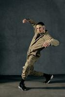 Athletic fellow with tattooed body and face, earrings, beard. Dressed in khaki jumpsuit, black sneakers. Dancing, gray background. Dancehall, hip-hop photo