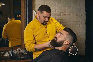 Client with big black beard during beard shaving in barber shop photo