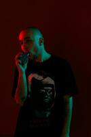 Close-up studio shot of a young bald guy vaping, blowing out a cloud of smoke on red background. photo