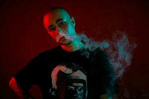 Close-up studio shot of a young bald guy vaping, blowing out a cloud of smoke on red background. photo