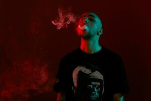 Close-up studio shot of a young bald guy vaping, blowing out a cloud of smoke on red background. photo