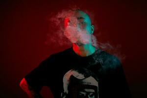 Close-up studio shot of a young bald guy vaping, blowing out a cloud of smoke on red background. photo