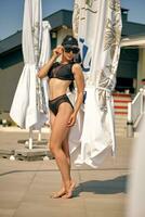 Portrait of a woman having rest and posing near a swimming pool. Dressed in a black swimsuit, sun visor and sunglasses. photo