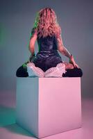 Young fitness blonde woman wearing blue velour booty shorts posing on camera, sits on top of big cube, back view photo