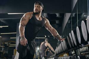 Young, tattooed male in black shorts, vest, cap. Going to do exercises with dumbbells for training his muscles in dark gym. Sport, fitness. Close up photo