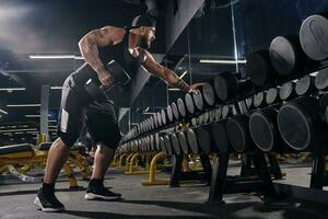 Tattooed, bearded, muscular sportsman in black shorts, vest, cap. Exercising with dumbbells for training his triceps, looking at the mirror. Close up photo