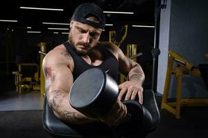 Tattooed, bearded guy in black vest and cap. He is lifting a dumbbell, training his biceps, sitting on preacher curl bench at dark gym. Close up photo