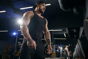 Tattooed male in black shorts, vest, cap. Going to do exercises with dumbbells, looking at mirror near set of black weights. Dark gym. Close up photo