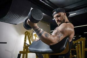 Tattooed guy with earring, in black vest and cap. He is lifting a dumbbell, training his biceps, sitting on preacher curl bench at dark gym. Close up photo