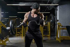 deportista en negro deporte guantes, bermudas, chaleco y gorra. él es levantamiento barra con pesas, formación su bíceps, posando en oscuro gimnasio con amarillo equipo. cerca arriba foto